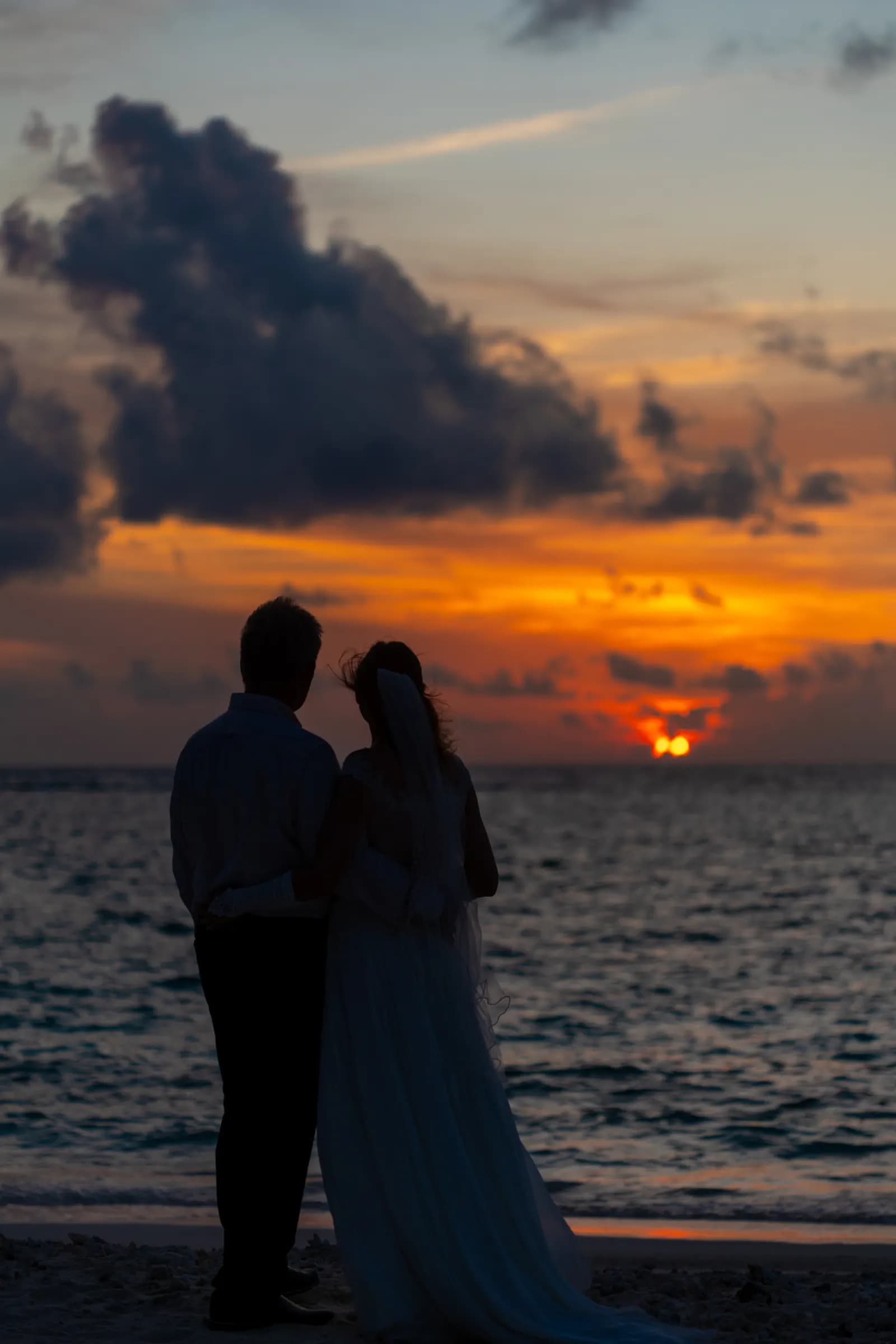 A couple on the beach at dusk, the sky pink behind them over flat water