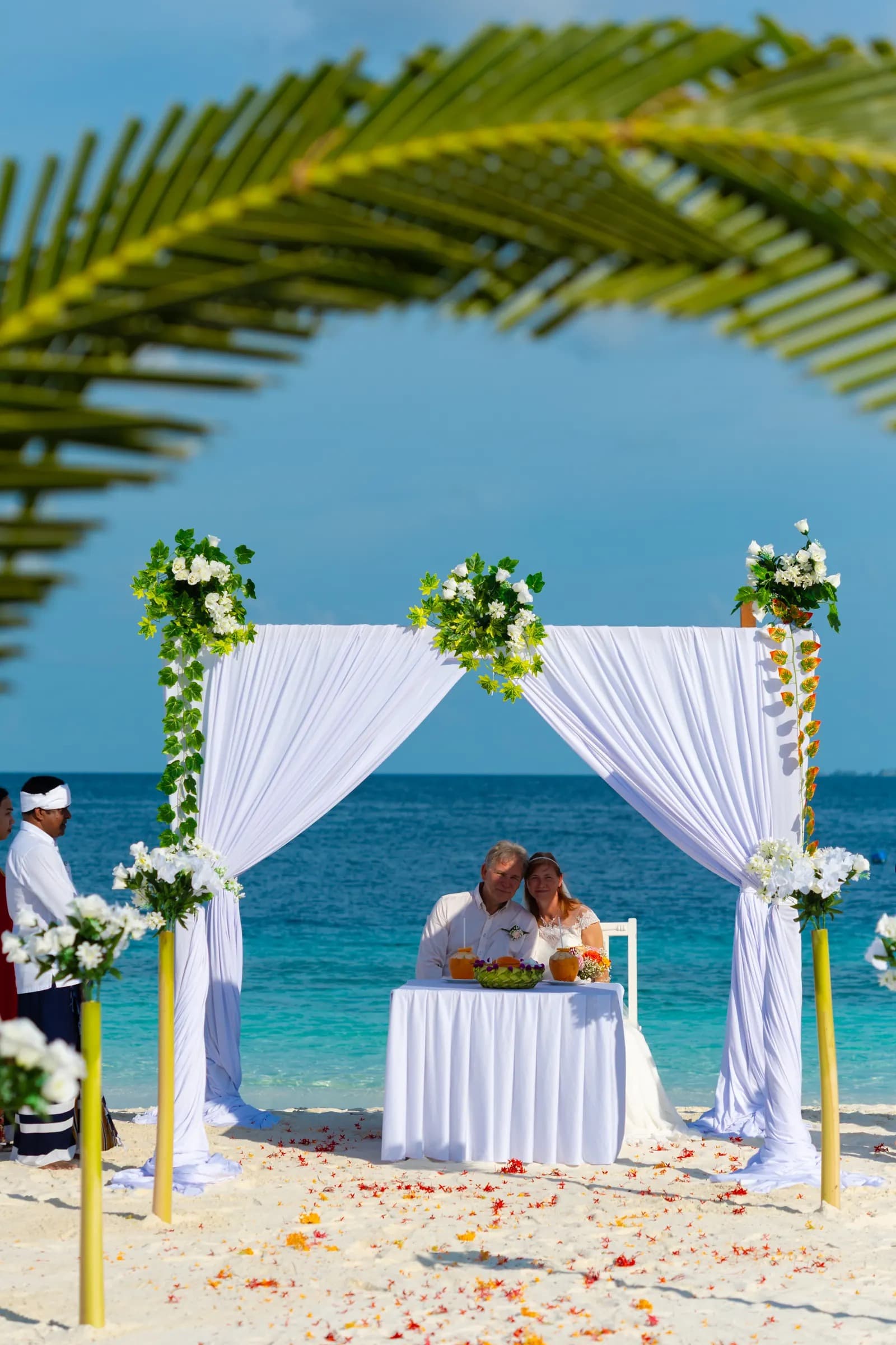 A couple seated under the white canopy, petals scattered along the sand in front of them